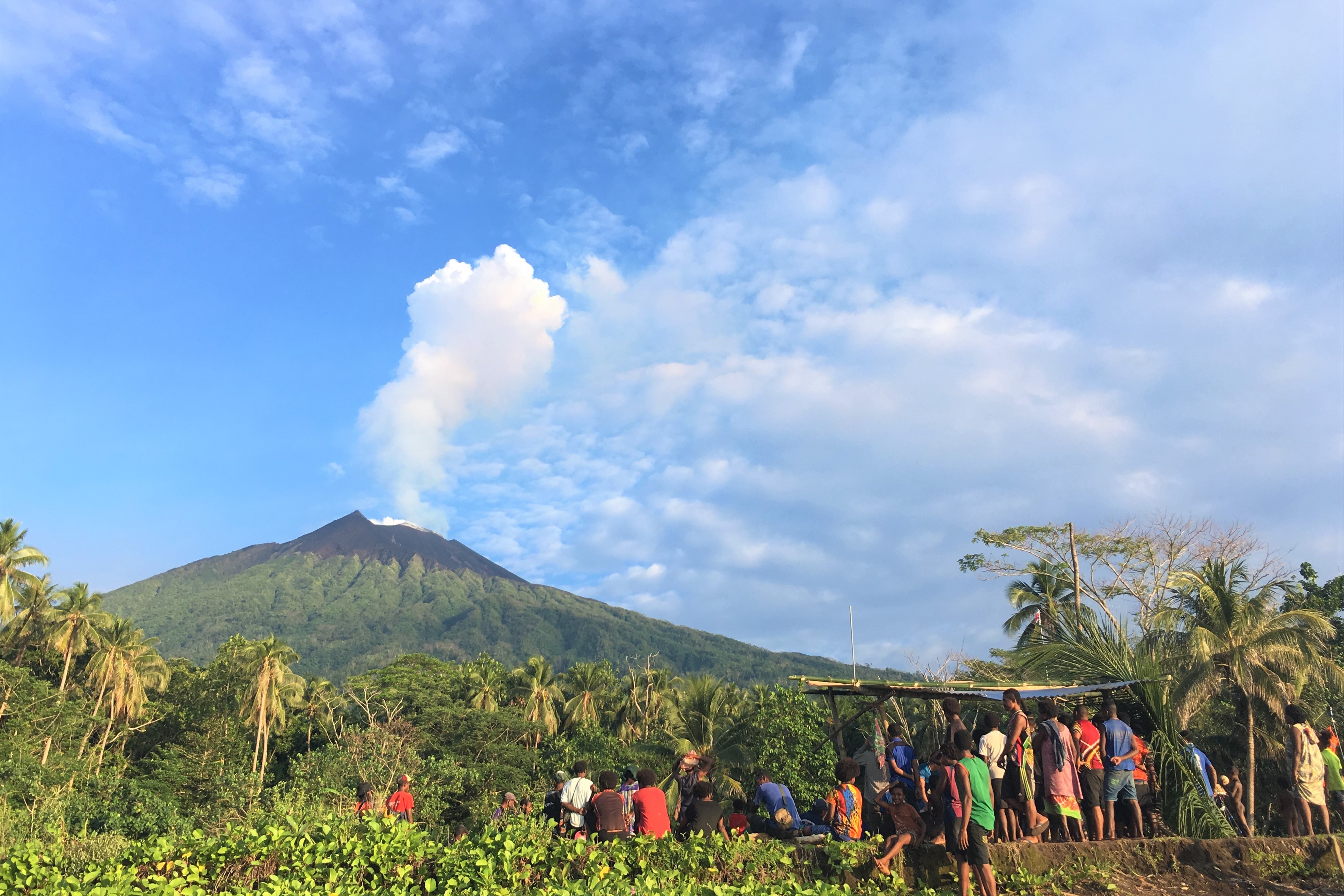 Manam islanders gathering together in the shadow of the volcano, which is showing a white gas plume against the bright blue sky; image credit Emma Liu, UCL