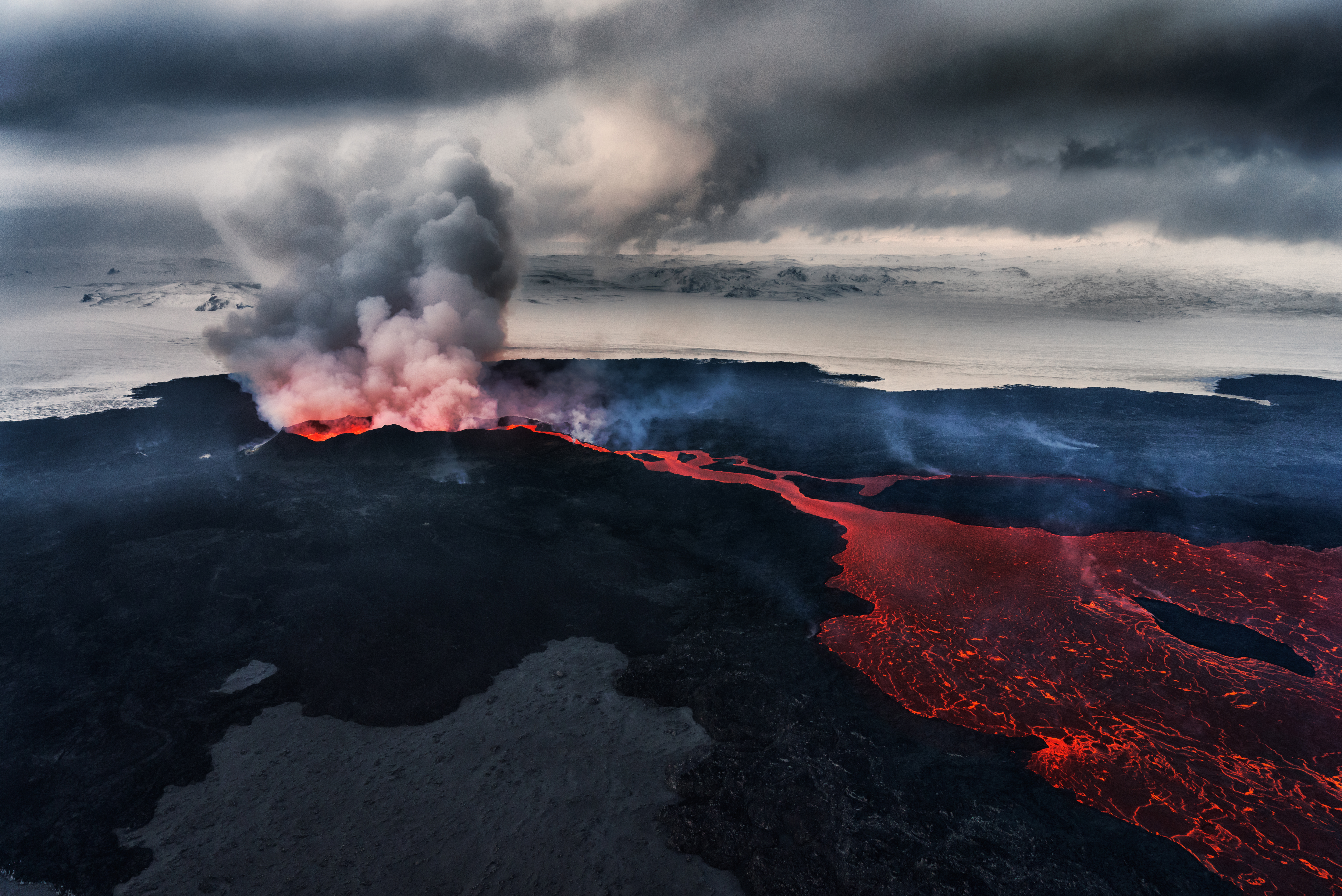 An aerial photo of the Holahraun fissure eruption, with a volcanic gas plume issuing from the vent and a lava flow extending from a mini volcanic cone across the sandur.