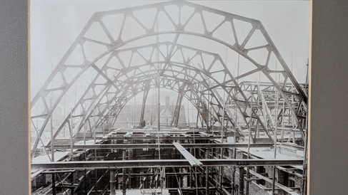 Black and white photo of building mid-construction, showing four arcing ribs of ironwork to support a roof.