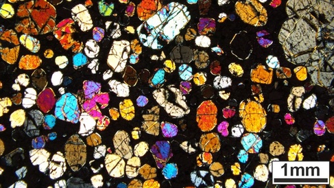 Thin section photo of rock, showing rounded colourful crystals ranging from yellow-orange to pink-blue. The background material is black.