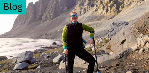 Photo of a woman stood on a beach, in front of a mountain, they are dressed in field gear and using crutches.