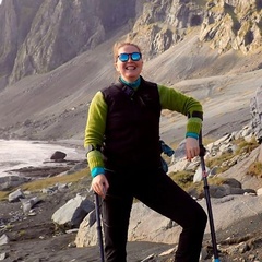 Photo of a woman stood on a beach, in front of a mountain, they are dressed in field gear and using crutches.