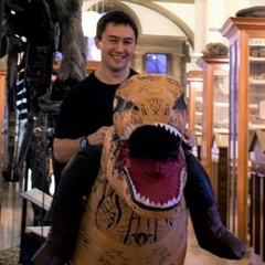 Image of a man smiling, wearing an inflatable dinosaur costume and stood inside the Sedgwick Museum
