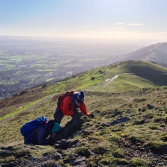 Photo of three scientists looking at a rock outcrop on top of the Malverns. It is a sunny day and the landscape stretches into the distance. 