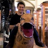 Image of a man smiling, wearing an inflatable dinosaur costume and stood inside the Sedgwick Museum