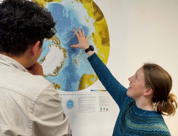 Photo of two people looking at a map of the Arctic, one is pointing above Greenland
