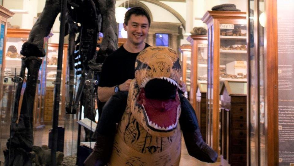 Image of a man smiling, wearing an inflatable dinosaur costume and stood inside the Sedgwick Museum