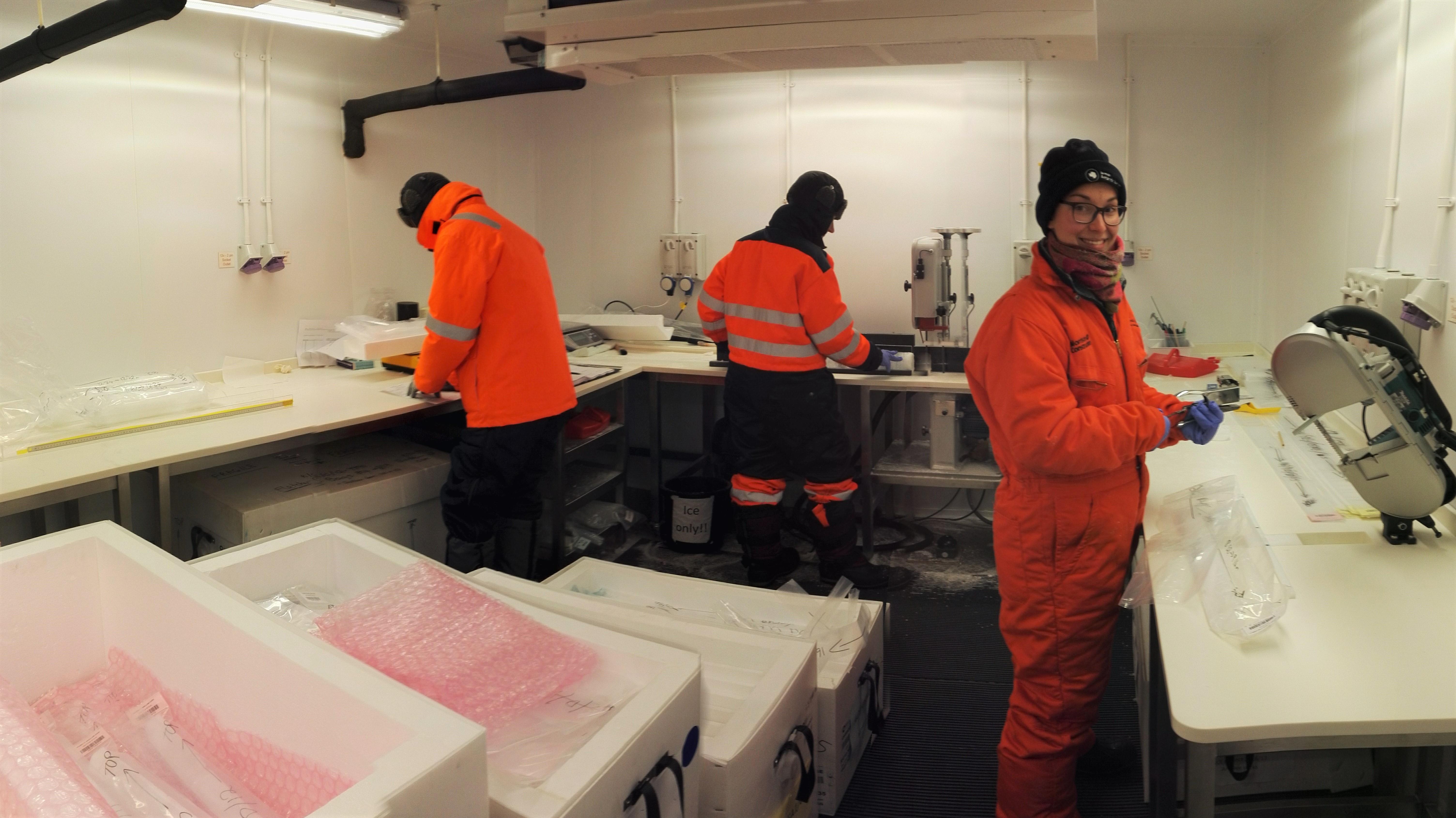 Dorethea processing ice core samples, credit: Amy King Image of Dorethea in the lab with two other scientists, they are wearing orange polar gear and sorting through ice cores