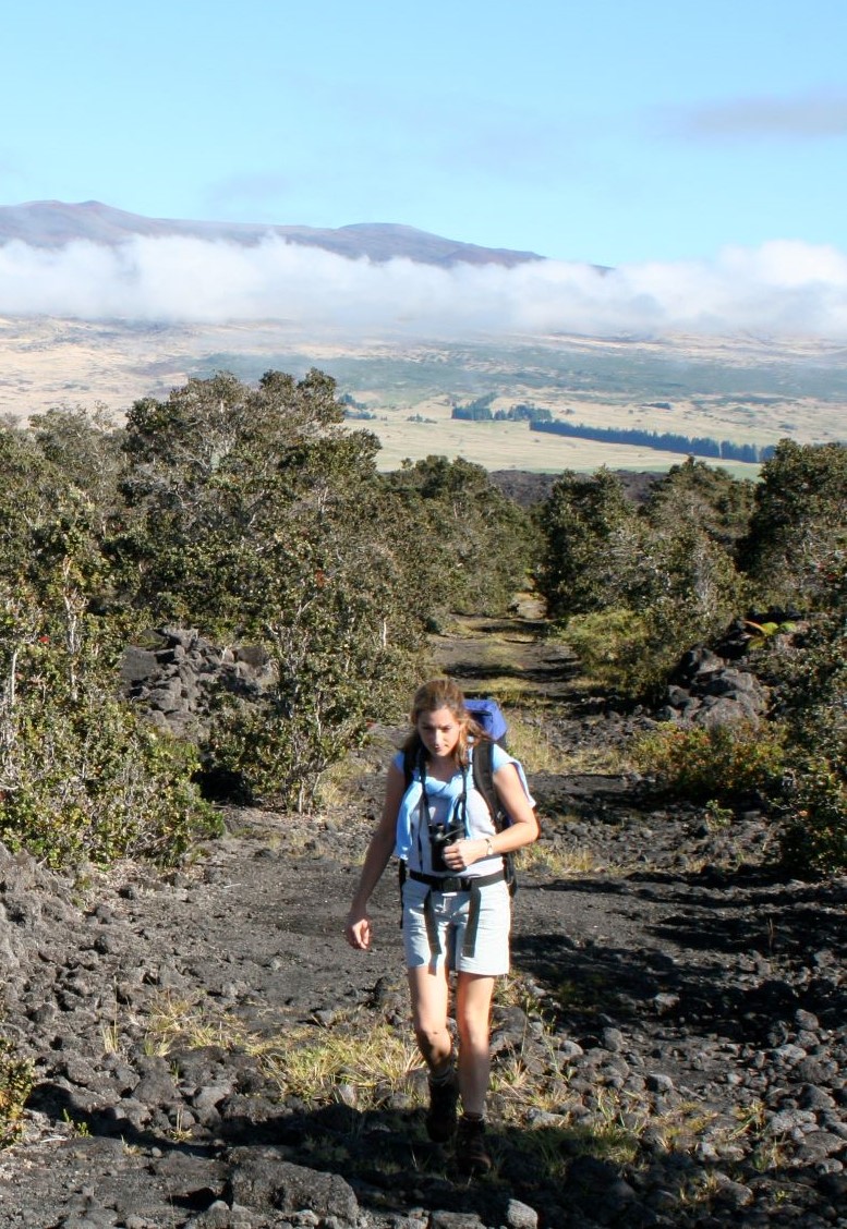 Image of Marie Edmonds walking up a scree covered incline, with the profile of Mauna Kea volcano in the background