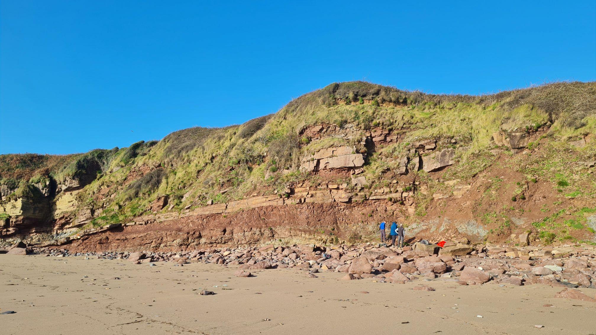 Photo of a sunny beach with three people work close to the red cliff face