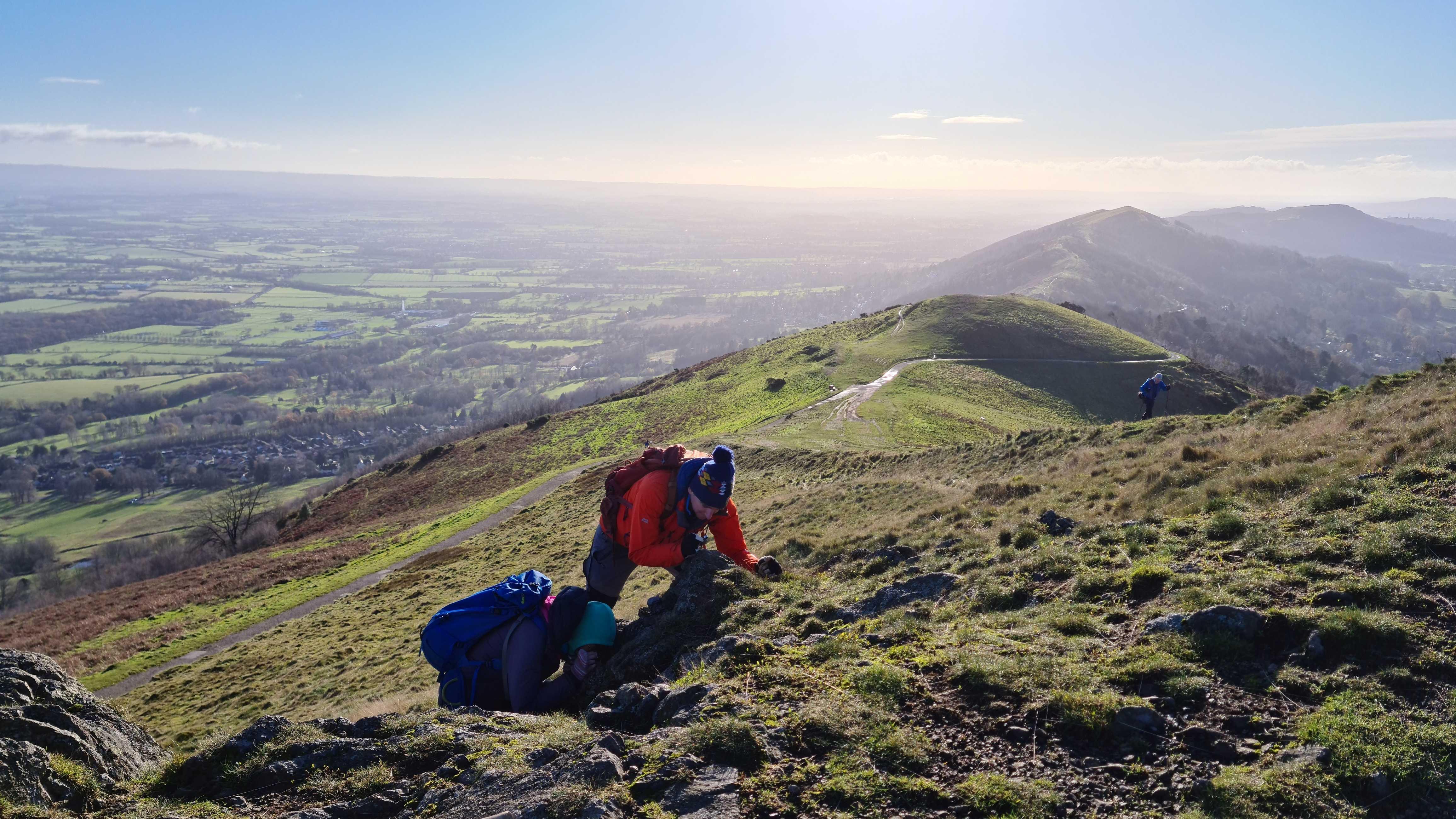 Photo of three scientists looking at a rock outcrop on top of the Malverns. It is a sunny day and the landscape stretches into the distance. 