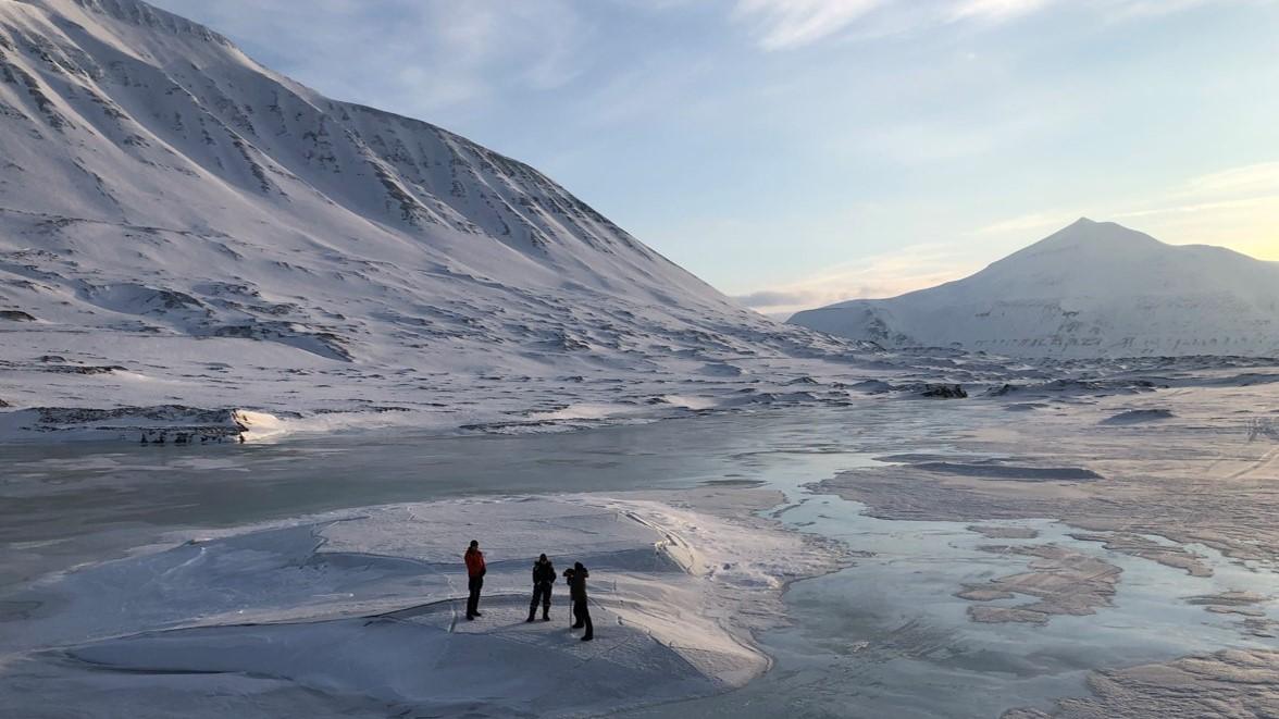 Photo of three people standing in front of a glacier, there are snowy mountains behind